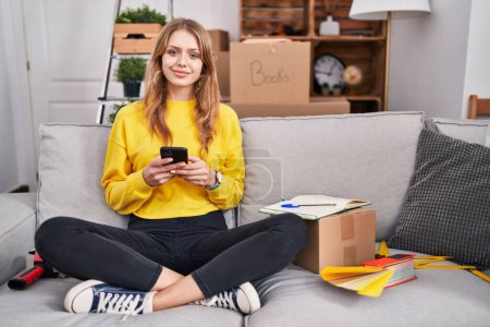Photo for Young blonde woman using smartphone sitting on sofa at new home - Royalty Free Image
