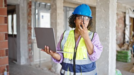 Photo for Young beautiful latin woman builder using laptop talking by walkie talkie at construction site - Royalty Free Image