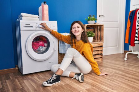 Téléchargez les photos : Jeune femme blonde assise sur le sol tournant la machine à laver à la buanderie - en image libre de droit