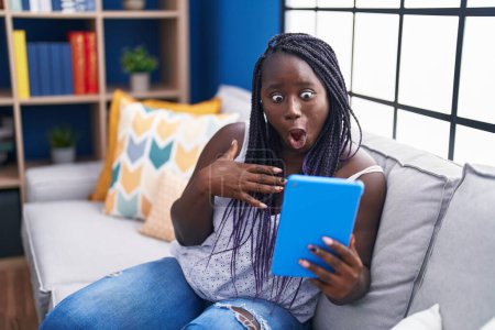 Foto de Mujer africana joven usando touchpad sentado en el sofá asustado y sorprendido con la boca abierta para sorpresa, cara de incredulidad - Imagen libre de derechos