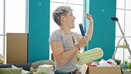 Photo for Young woman singing song using paint roller with microphone dancing at new home - Royalty Free Image