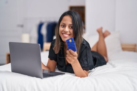 Photo for Young hispanic woman using laptop and credit card lying on bed at bedroom - Royalty Free Image