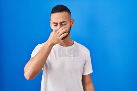 Foto de Young hispanic man standing over blue background smelling something stinky and disgusting, intolerable smell, holding breath with fingers on nose. bad smell - Imagen libre de derechos