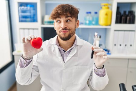 Téléchargez les photos : Homme arabe avec barbe travaillant au laboratoire scientifique tenant des échantillons de sang souriant regardant vers le côté et regardant loin en pensant. - en image libre de droit