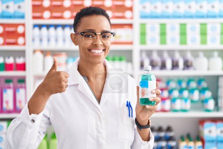 Téléchargez les photos : Femme afro-américaine travaillant à la pharmacie pharmacie tenant sirop souriant heureux et positif, pouce levé faire excellent et signe d'approbation - en image libre de droit