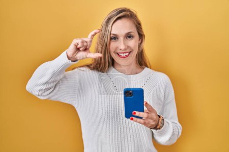 Photo for Young blonde woman using smartphone typing message smiling and confident gesturing with hand doing small size sign with fingers looking and the camera. measure concept. - Royalty Free Image