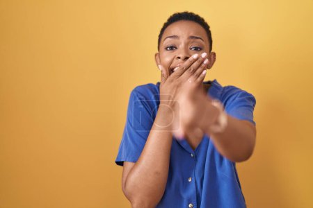 Foto de Mujer afroamericana de pie sobre fondo amarillo riéndose de ti, señalando con el dedo a la cámara con la mano sobre la boca, expresión de vergüenza - Imagen libre de derechos