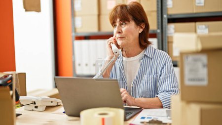 Téléchargez les photos : Femme d'âge moyen e-commerce travailleur d'affaires en utilisant un ordinateur portable parlant au téléphone au bureau - en image libre de droit
