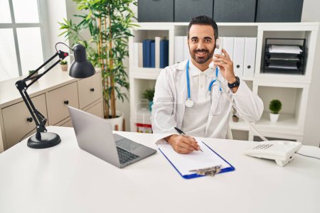 Foto de Un joven hispano vistiendo uniforme médico hablando por teléfono en la clínica - Imagen libre de derechos