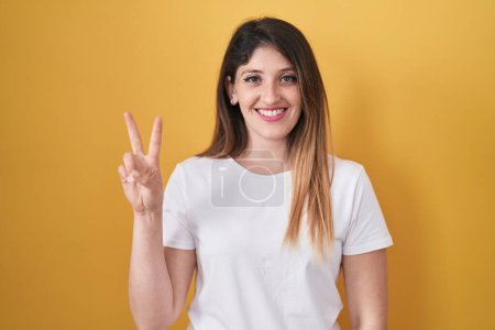 Téléchargez les photos : Jeune femme brune debout sur fond jaune montrant et pointant vers le haut avec les doigts numéro deux tout en souriant confiant et heureux. - en image libre de droit