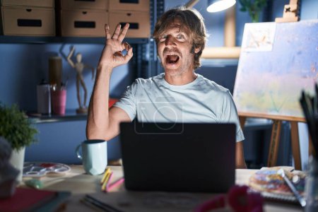 Téléchargez les photos : Homme d'âge moyen assis au studio d'art avec ordinateur portable la nuit souriant positif faire ok signe avec la main et les doigts. expression réussie. - en image libre de droit