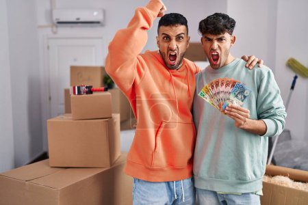 Foto de Joven pareja gay hispana mudándose a una nueva casa sosteniendo billetes molestos y frustrados gritando de rabia, gritando loca de rabia y levantando la mano - Imagen libre de derechos