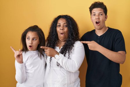 Photo for Family of mother, daughter and son standing over yellow background surprised pointing with finger to the side, open mouth amazed expression. - Royalty Free Image