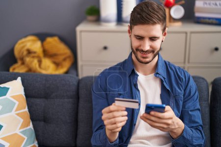 Téléchargez les photos : Young caucasian man using smartphone and credit card sitting on sofa at home - en image libre de droit
