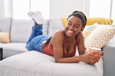 Téléchargez les photos : Femme afro-américaine buvant du café allongé sur le canapé à la maison - en image libre de droit