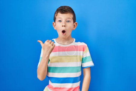 Photo for Young caucasian kid standing over blue background surprised pointing with hand finger to the side, open mouth amazed expression. - Royalty Free Image