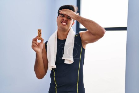 Photo for Hispanic man eating protein bar as healthy energy snack stressed and frustrated with hand on head, surprised and angry face - Royalty Free Image