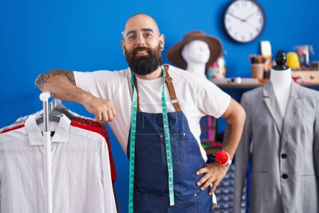 Photo for Young bald man tailor smiling confident leaning on clothes rack at clothing factory - Royalty Free Image