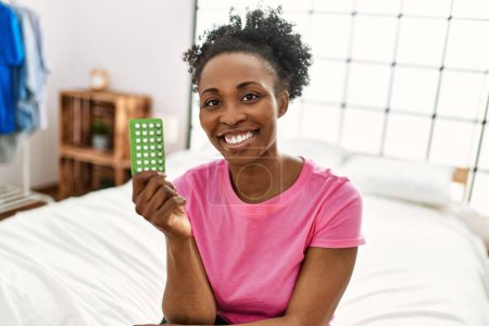 Photo for African american woman holding birth control pills sitting on bed at bedroom - Royalty Free Image