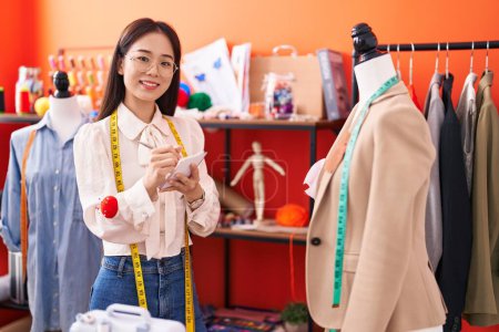 Téléchargez les photos : Jeune femme chinoise tailleur écriture sur carnet regardant veste à l'atelier - en image libre de droit