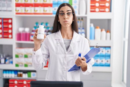 Téléchargez les photos : Jeune femme brune travaillant à la pharmacie drugstore tenant des pilules gonflant les joues avec un visage drôle. bouche gonflée d'air, attrapant l'air. - en image libre de droit