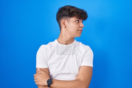 Téléchargez les photos : Adolescent hispanique debout sur fond bleu regardant du côté avec les bras croisés convaincu et confiant - en image libre de droit