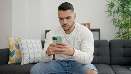 Photo for Young hispanic man stressed sitting on sofa using smartphone at home - Royalty Free Image