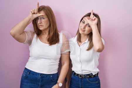 Photo for Hispanic mother and daughter wearing casual white t shirt over pink background making fun of people with fingers on forehead doing loser gesture mocking and insulting. - Royalty Free Image