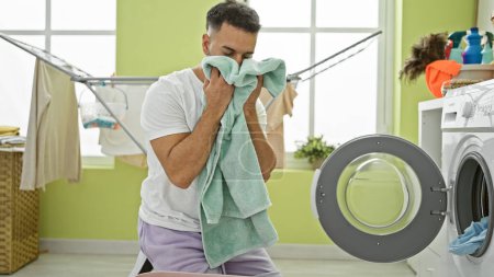 Photo for Young arab man washing clothes smelling clean towel at laundry room - Royalty Free Image