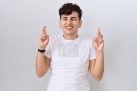 Téléchargez les photos : Jeune homme non binaire portant un t-shirt blanc décontracté geste doigt croisé sourire avec espoir et les yeux fermés. chance et concept superstitieux. - en image libre de droit