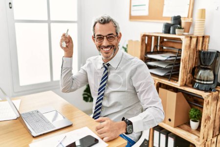 Foto de Hombre de pelo gris de mediana edad agente de bienes raíces con llave de nuevo hogar en la oficina - Imagen libre de derechos