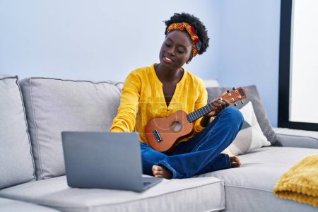 Foto de Joven africana americana mujer teniendo en línea ukelele clase sentado en sofá en casa - Imagen libre de derechos