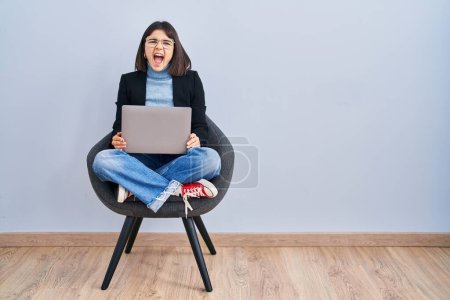 Téléchargez les photos : Jeune femme hispanique assis sur la chaise en utilisant ordinateur portable en colère et fou hurlant frustré et furieux, criant de colère. rage et concept agressif. - en image libre de droit