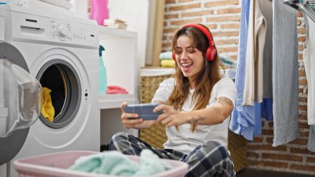 Téléchargez les photos : Jeune belle femme hispanique jouant jeu vidéo laver les vêtements à la buanderie - en image libre de droit