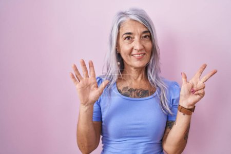 Foto de Mujer de mediana edad con tatuajes de pie sobre fondo rosa mostrando y apuntando hacia arriba con los dedos número ocho mientras sonríe confiado y feliz. - Imagen libre de derechos