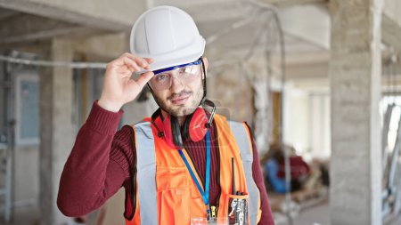 Téléchargez les photos : Jeune homme hispanique constructeur debout avec un visage sérieux sur le chantier de construction - en image libre de droit
