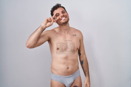 Téléchargez les photos : Jeune homme hispanique debout torse nu portant des sous-vêtements faisant symbole de paix avec les doigts sur le visage, souriant joyeux montrant la victoire - en image libre de droit