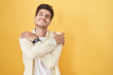 Foto de Joven hombre hispano de pie sobre fondo amarillo abrazándose feliz y positivo, sonriendo confiado. amor propio y cuidado personal - Imagen libre de derechos