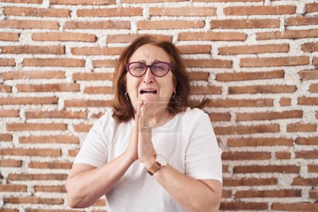 Photo for Senior woman with glasses standing over bricks wall begging and praying with hands together with hope expression on face very emotional and worried. begging. - Royalty Free Image