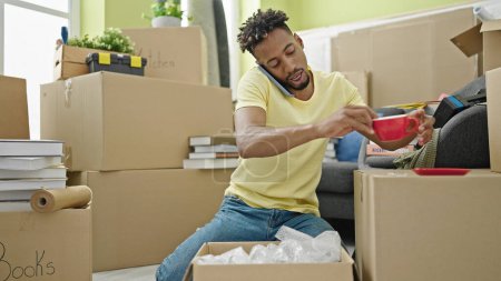 Photo for African american man unpacking cardboard box talking on smartphone at new home - Royalty Free Image
