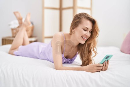 Téléchargez les photos : Jeune femme blonde utilisant un smartphone couché sur le lit dans la chambre - en image libre de droit
