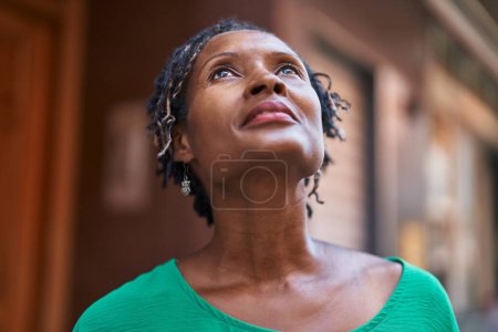 Téléchargez les photos : Moyen Age femme afro-américaine regardant le ciel avec une expression sérieuse dans la rue - en image libre de droit