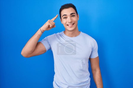 Photo for Young hispanic man standing over blue background smiling pointing to head with one finger, great idea or thought, good memory - Royalty Free Image