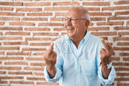 Foto de Senior man with grey hair standing over bricks wall showing middle finger doing fuck you bad expression, provocation and rude attitude. screaming excited - Imagen libre de derechos