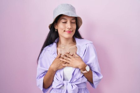 Téléchargez les photos : Jeune femme hispanique debout sur fond rose portant un chapeau souriant avec les mains sur la poitrine avec les yeux fermés et geste reconnaissant sur le visage. concept de santé. - en image libre de droit