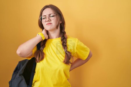 Téléchargez les photos : Jeune femme caucasienne portant un sac à dos étudiant sur fond jaune souffrant d'une blessure au cou, touchant le cou avec la main, douleur musculaire - en image libre de droit