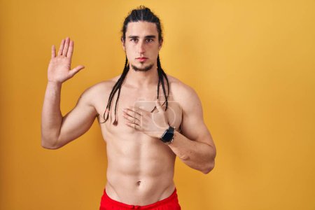 Téléchargez les photos : Homme hispanique aux cheveux longs, torse nu sur fond jaune, jurant la main sur la poitrine et la paume ouverte, prêtant serment de fidélité - en image libre de droit