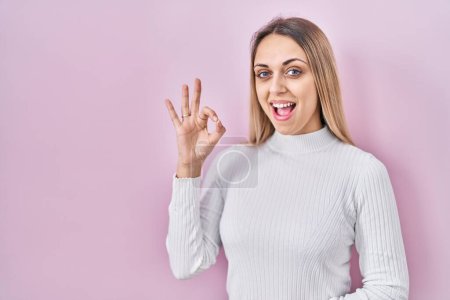 Téléchargez les photos : Jeune femme blonde portant un pull blanc sur fond rose souriant positif faisant signe ok avec la main et les doigts. expression réussie. - en image libre de droit