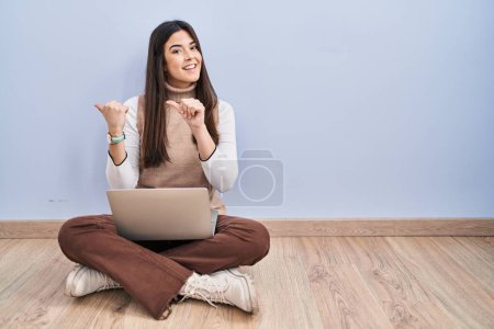 Foto de Mujer morena joven trabajando con computadora portátil sentada en el suelo apuntando a la espalda con la mano y los pulgares hacia arriba, sonriendo confiado - Imagen libre de derechos