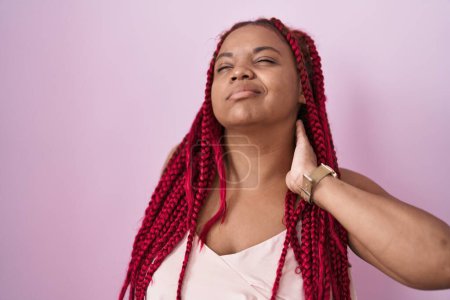 Foto de Mujer afroamericana con cabello trenzado de pie sobre fondo rosa sufriendo de dolor de cuello lesión, tocando el cuello con la mano, dolor muscular - Imagen libre de derechos